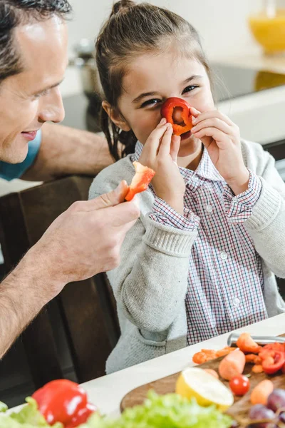 Heureux père avec fille, petit enfant applique tranche de poivre au nez et en regardant la caméra à la cuisine — Photo de stock