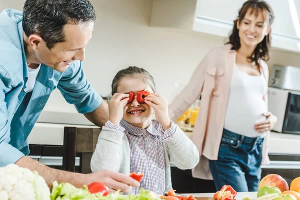 Famille heureuse, petit enfant applique des tranches de poivre aux yeux lorsque le père et la mère enceinte la regardent à la cuisine — Photo de stock