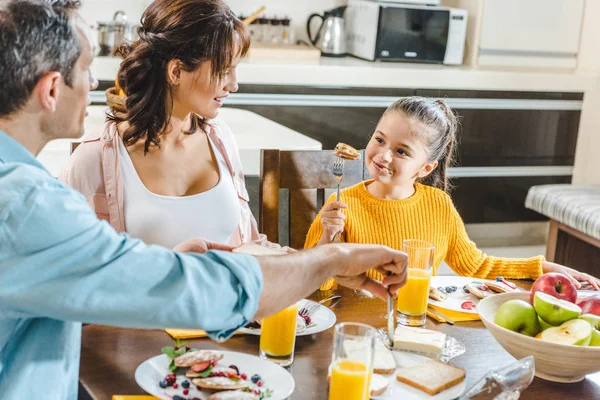 Família feliz comer panquecas à mesa com suco e frutas na cozinha — Fotografia de Stock