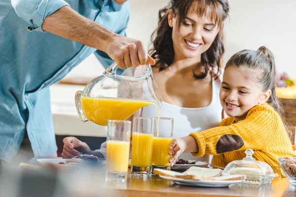 Famille heureuse à table, homme versant du jus dans des verres à la femme et l'enfant à la cuisine — Photo de stock