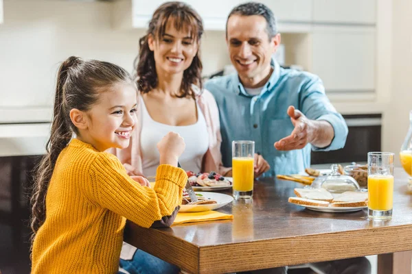 Alegre familia sentada a la mesa con panqueques, padre señalando el jugo a mano en la cocina - foto de stock