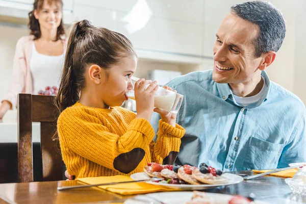 Счастливая семья на кухне, дочь с отцом сидит за столом с блинами и смотрит друг на друга, когда дочь пьет молоко — стоковое фото