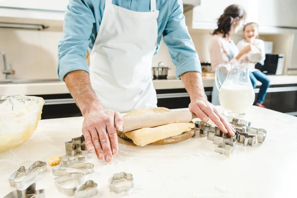 Cropped image of man rolling out dough by rolling pin on table in front of mother with daughter at kitchen — Stock Photo