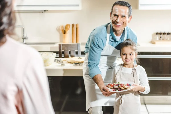 Feliz padre con hija mostrando plato con panqueques a la madre en la cocina - foto de stock