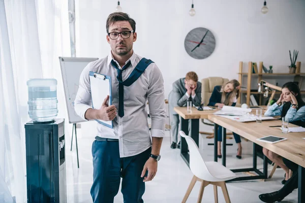 Overworked untidy businessman with clipboard standing at office while colleagues sitting at conference hall — Stock Photo