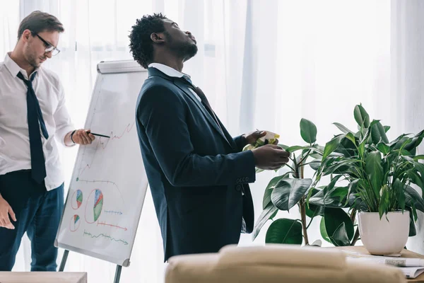 Sleepy african american manager spraying plant with water at office while his colleague making presentation — Stock Photo