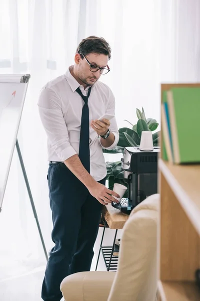 Untidy businessman pouring coffee from office machine and using smartphone — Stock Photo