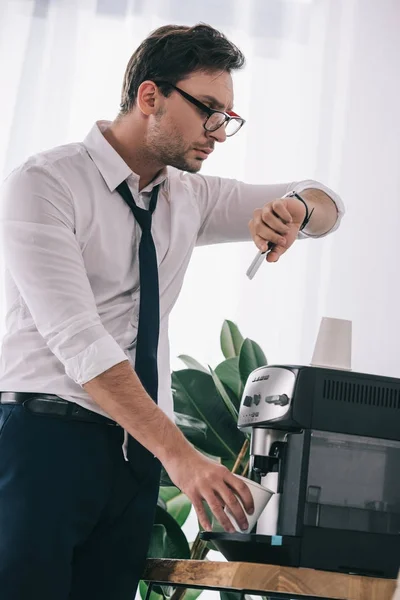 Hombre de negocios vertiendo café de la máquina de oficina y mirando el reloj — Stock Photo