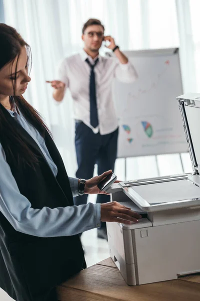 Jeune gestionnaire utilisant copieur au bureau tandis que son collègue parlant par téléphone — Photo de stock