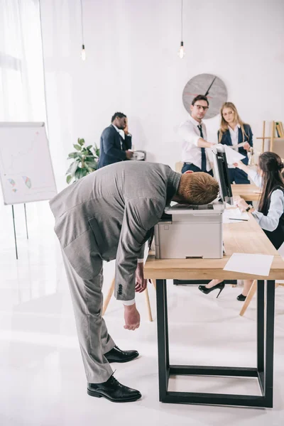 Exhausted businessman sleeping on copier at office while colleagues having conversation — Stock Photo