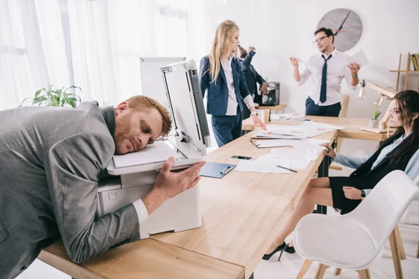 Erschöpfter Zombie wie Geschäftsmann, der auf Kopierer schläft, während seine Kollegen im Büro Gespräche führen — Stockfoto