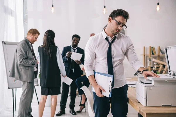 Young handsome businessman using copier at office with blurred colleagues on background — Stock Photo