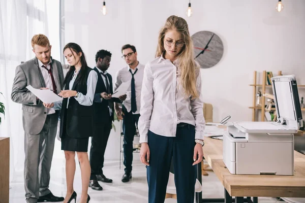 Femme d'affaires épuisée debout près copieur avec des collègues travaillant sur fond au bureau — Photo de stock