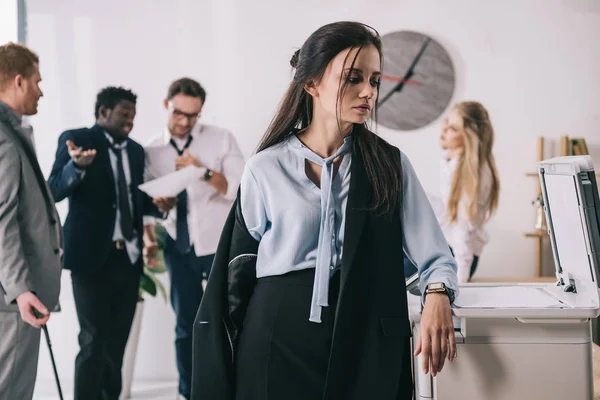 Sovraccarico di lavoro donna d'affari disordinata in piedi vicino copiatrice con colleghi sfocati che lavorano su sfondo in ufficio — Foto stock