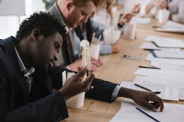 Exhausted african american businessman eating noodles with his colleagues at office — Stock Photo