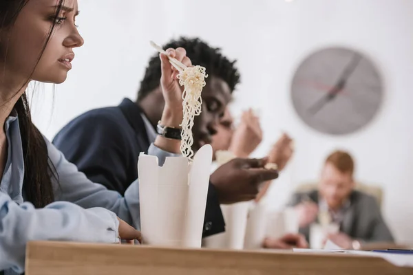 Exhausted young businesswoman eating noodles with colleagues at office — Stock Photo