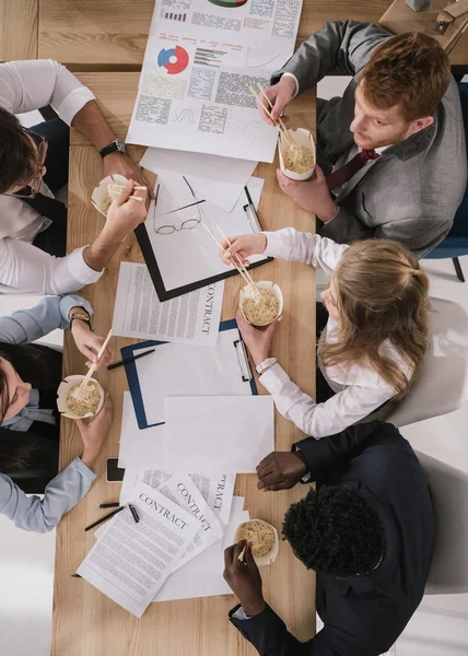 Von oben: Geschäftsleute essen Nudeln, während sie im Büro Papierkram erledigen — Stockfoto