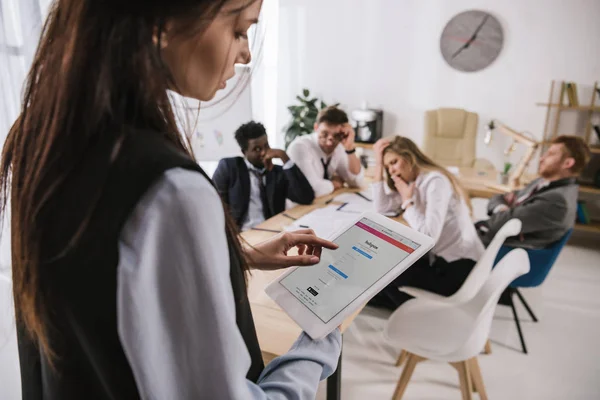 Empresaria usando tableta con aplicación instagram en la pantalla en la oficina con colegas desenfocados y sobrecargados de trabajo sentados en la sala de conferencias - foto de stock