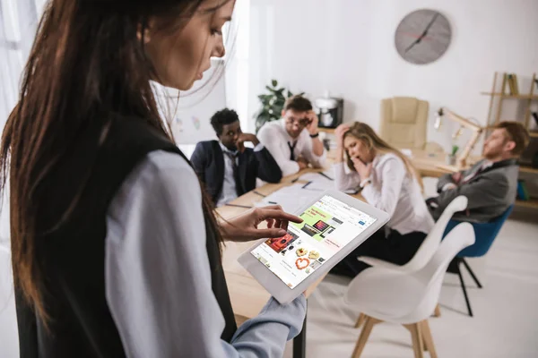 Joven mujer de negocios utilizando la tableta con el sitio web de eBay en la pantalla, mientras que los colegas tienen conversación - foto de stock