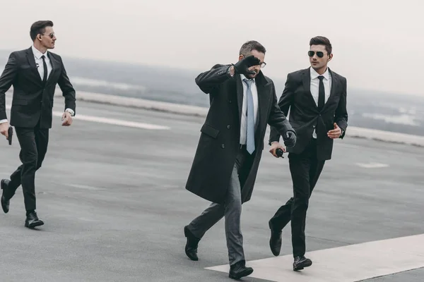 Hombre de negocios cubriéndose la cara con la mano y caminando con guardaespaldas en helipuerto - foto de stock