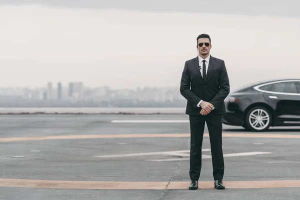Serious bodyguard standing with sunglasses and security earpiece on helipad and looking at camera — Stock Photo