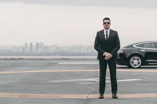 Garde du corps sérieux debout avec des lunettes de soleil et un écouteur de sécurité sur l'héliport et regardant la caméra — Photo de stock