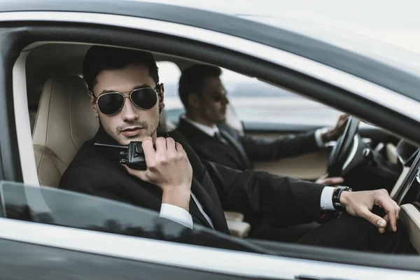 Bodyguard in sunglasses talking by portable radio while sitting in car — Stock Photo