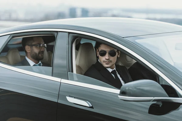 Dos guardaespaldas y hombre de negocios en gafas sentados en un coche negro - foto de stock