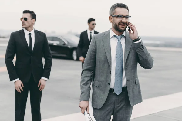 Uomo d'affari sorridente che cammina con guardie del corpo e parla con lo smartphone — Foto stock