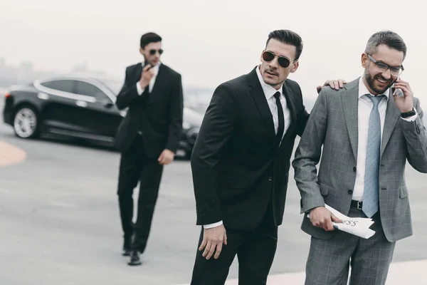 Happy businessman walking with bodyguards and talking by smartphone — Stock Photo