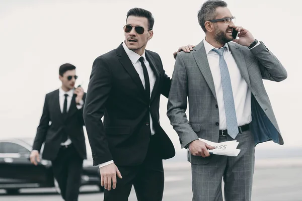 Hombre de negocios caminando con dos guardaespaldas y hablando por teléfono inteligente - foto de stock