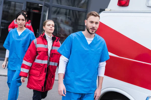 Jovens paramédicos masculinos e femininos caminhando perto de ambulância — Fotografia de Stock