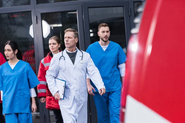 Team of serious paramedics with doctor going from hospital to ambulance — Stock Photo