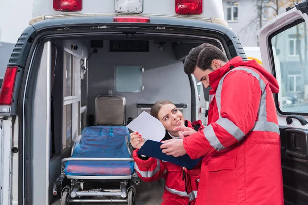 Paramédicos hombres y mujeres felices leyendo documentos delante de la ambulancia - foto de stock