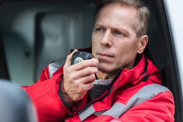 Handsome middle aged male paramedic talking by portable radio while sitting in ambulance — Stock Photo