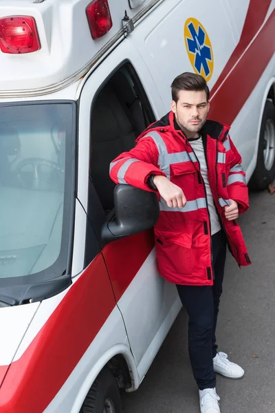 Grave joven paramédico masculino de pie y apoyado en la ambulancia - foto de stock