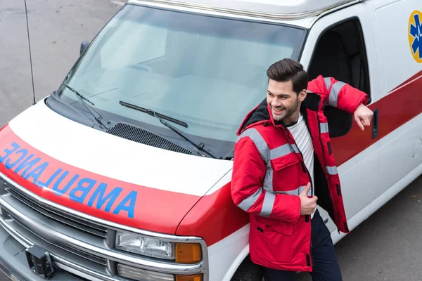 Sonriente guapo médico masculino de pie y apoyándose en la ambulancia - foto de stock