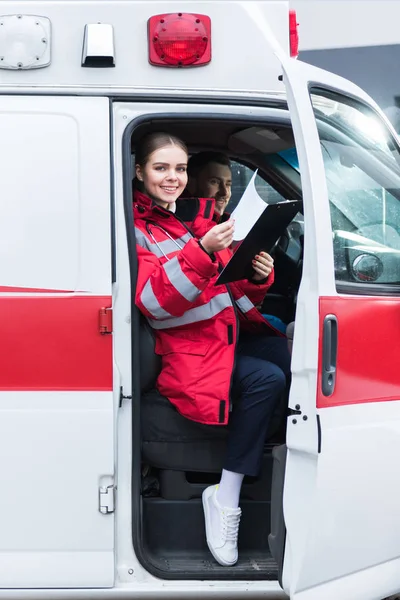 Smiling paramedic sitting in ambulance and holding clipboard — Stock Photo