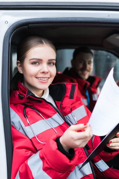 Smiling young female paramedic holding clipboard and looking at camera — Stock Photo