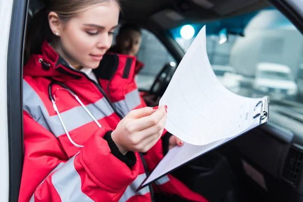 Paramédico femenino sentado en ambulancia y mirando el portapapeles - foto de stock