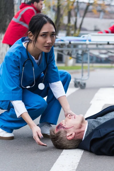 Young female Asian doctor helping unconscious injured man lying on a street — Stock Photo