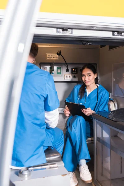 Sonriente asiático paramédico celebración portapapeles y sentado en ambulancia - foto de stock