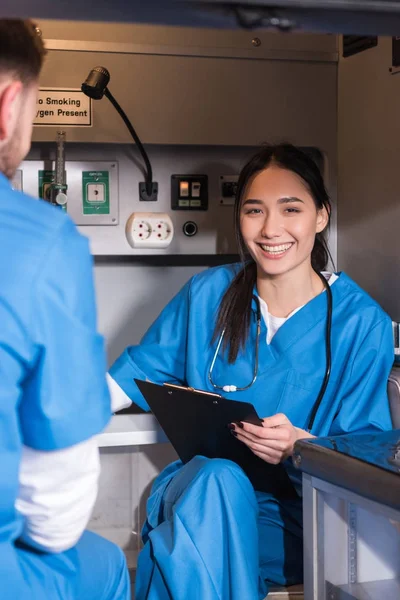 Sonriente asiático paramédico sentado en ambulancia y mirando a la cámara - foto de stock