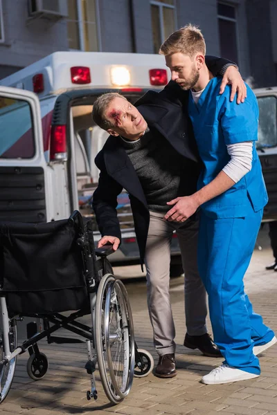 Paramédico ayudando al paciente lesionado a sentarse en silla de ruedas - foto de stock