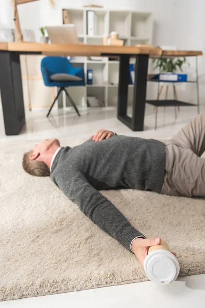 Hombre inconsciente acostado en un piso en la oficina y sosteniendo la taza de café desechable en la mano - foto de stock