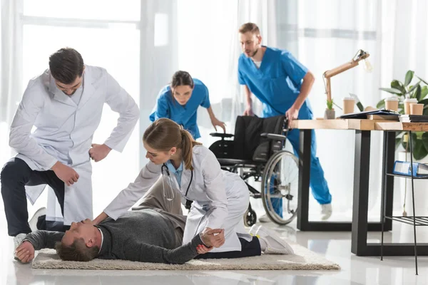 Los médicos jóvenes que comprueban el pulso del hombre inconsciente acostado en el suelo en el hospital - foto de stock