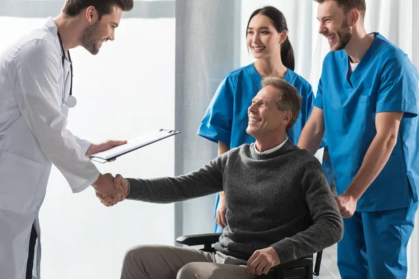 Sonriente médico y paciente en silla de ruedas estrechando las manos - foto de stock