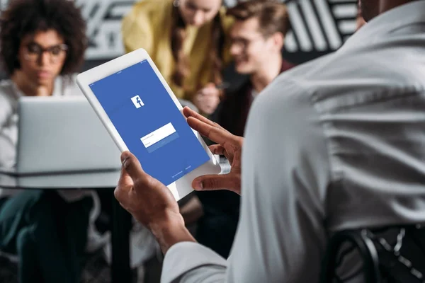 Homme utilisant tablette avec application facebook à l'écran avec ses partenaires d'affaires en arrière-plan — Photo de stock
