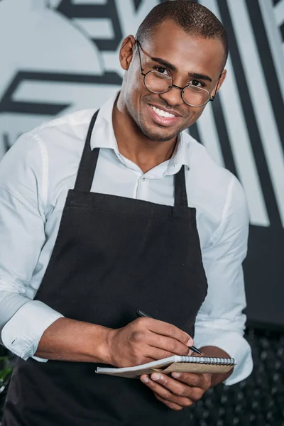 Beau jeune serveur écrit dans le bloc-notes et en regardant la caméra — Photo de stock