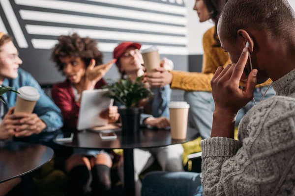 Young man listening music with earphones while his friends blurred sitting on background — Stock Photo